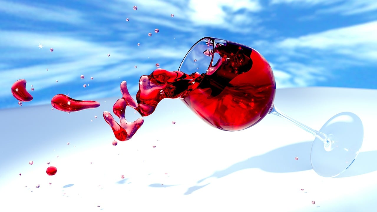 A wine glass tipped on its side, red wine splashing out against a bright blue sky